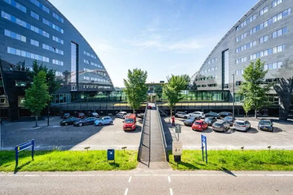 Moderne kantoorgebouwen aan de Rivium Promenade met een parkeergarage en voetgangersbrug op een zonnige dag.
